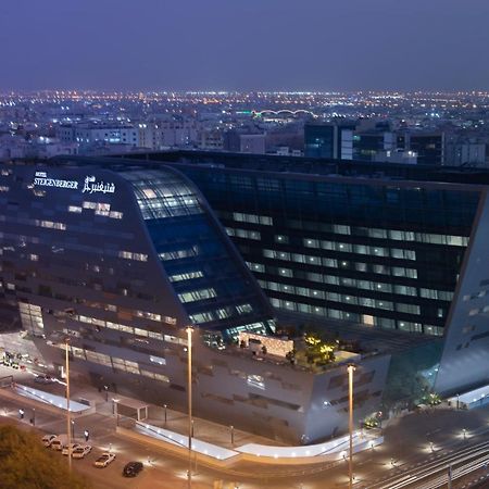 Steigenberger Hotel Доха Екстер'єр фото The image shows a modern architectural building, likely a hotel or office structure, illuminated at night. The design features a unique angular shape with large glass windows, reflecting the surrounding city lights. The building appears to be part of