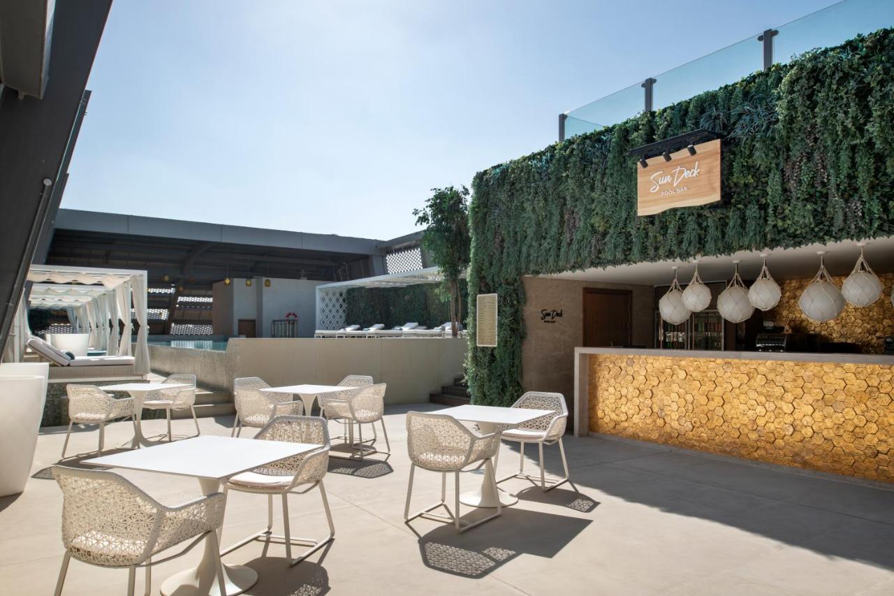 Steigenberger Hotel Доха Екстер'єр фото The photo shows a stylish outdoor seating area of a modern venue, likely a restaurant or bar. The space features several white tables and chairs arranged neatly on a light-colored floor. In the background, there's a bar area with a warm-toned wood fa