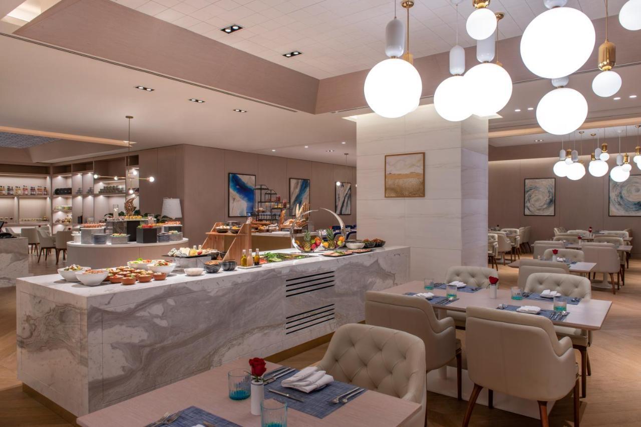 Steigenberger Hotel Доха Екстер'єр фото The photo shows a modern restaurant interior that features a stylish and elegant design. In the foreground, there are tables with light-colored upholstered chairs, set with blue napkins and small flower arrangements. 

In the background, there is a b