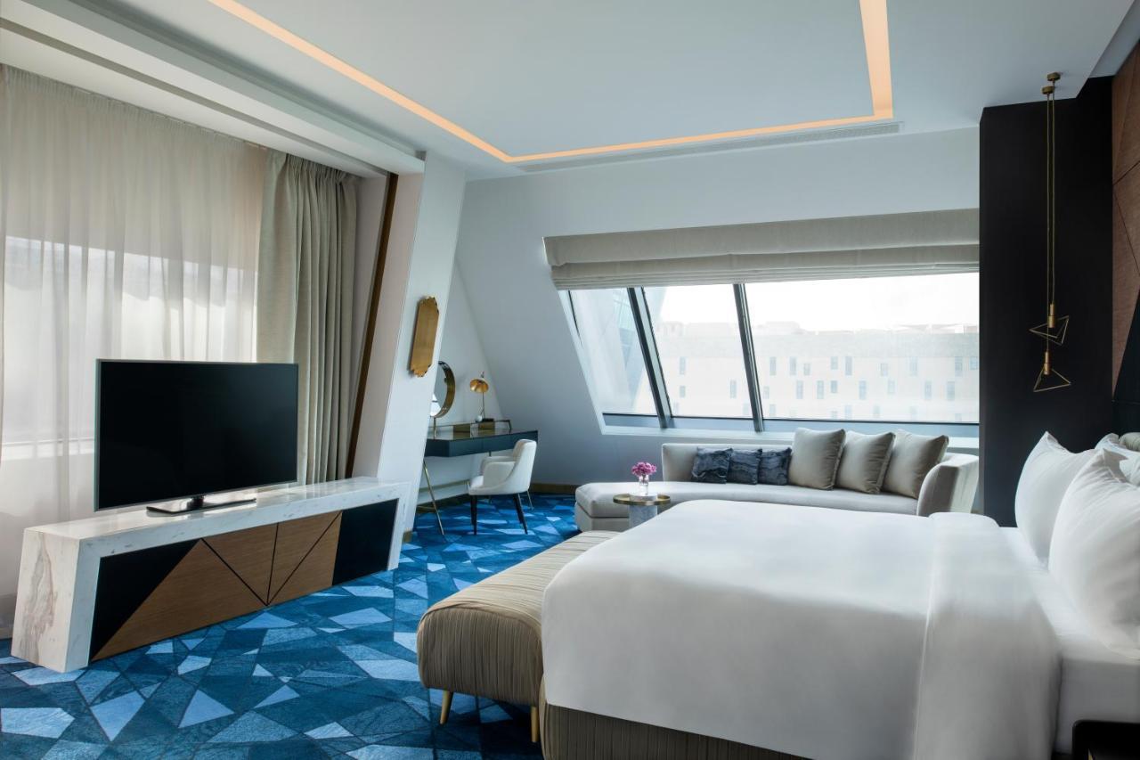 Steigenberger Hotel Доха Екстер'єр фото The photo shows a modern hotel room featuring a sleek design. The room includes a large bed with white linens, a stylish seating area with gray and beige tones, and a flat-screen television mounted on a wall. There are large windows that allow natura