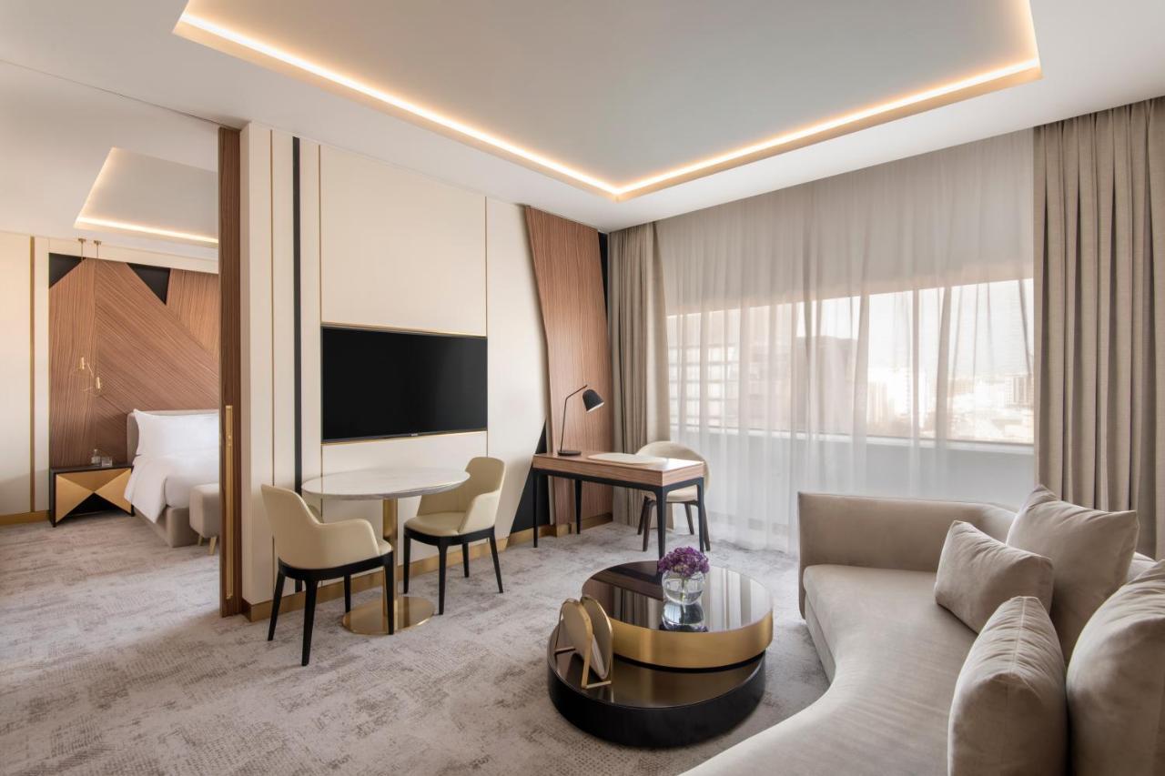 Steigenberger Hotel Доха Екстер'єр фото The photo shows a modern hotel room that features a stylish and contemporary design. There is a comfortable seating area with a light-colored sofa and a round coffee table, along with two chairs and a small desk. A large flat-screen television is mou