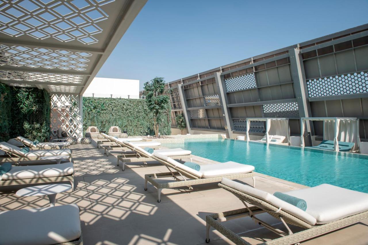 Steigenberger Hotel Доха Екстер'єр фото The photo shows a modern outdoor pool area. There are several lounge chairs arranged around the pool, which is a vibrant blue color. Sunlight casts intricate shadows on the ground due to a lattice structure overhead. On one side, there are green plan