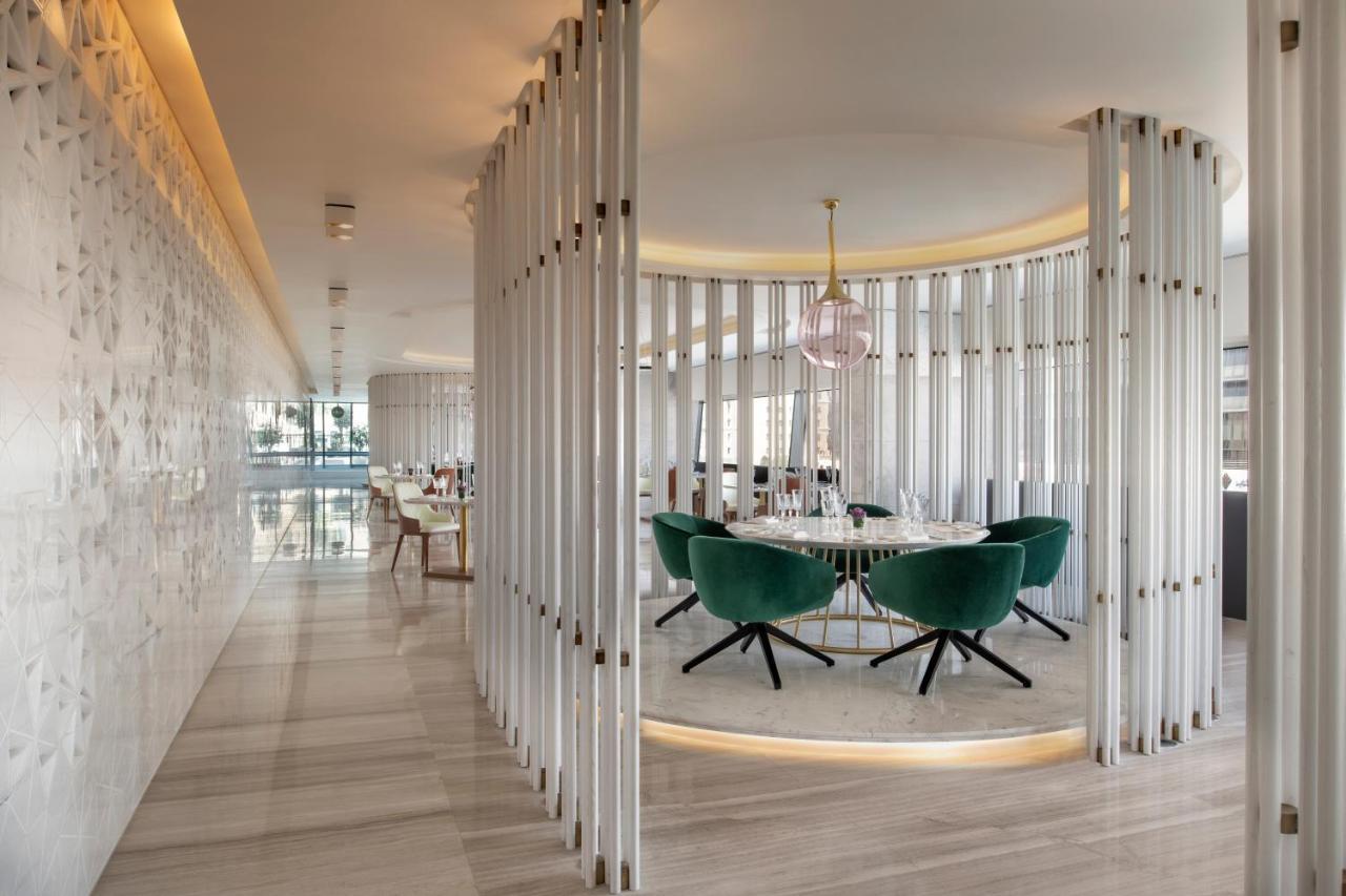 Steigenberger Hotel Доха Екстер'єр фото The photo shows a modern, stylish interior of a restaurant or dining area. There is a circular dining table surrounded by green upholstered chairs with a sleek design. The space is framed by vertical white panels, creating a sense of separation and p