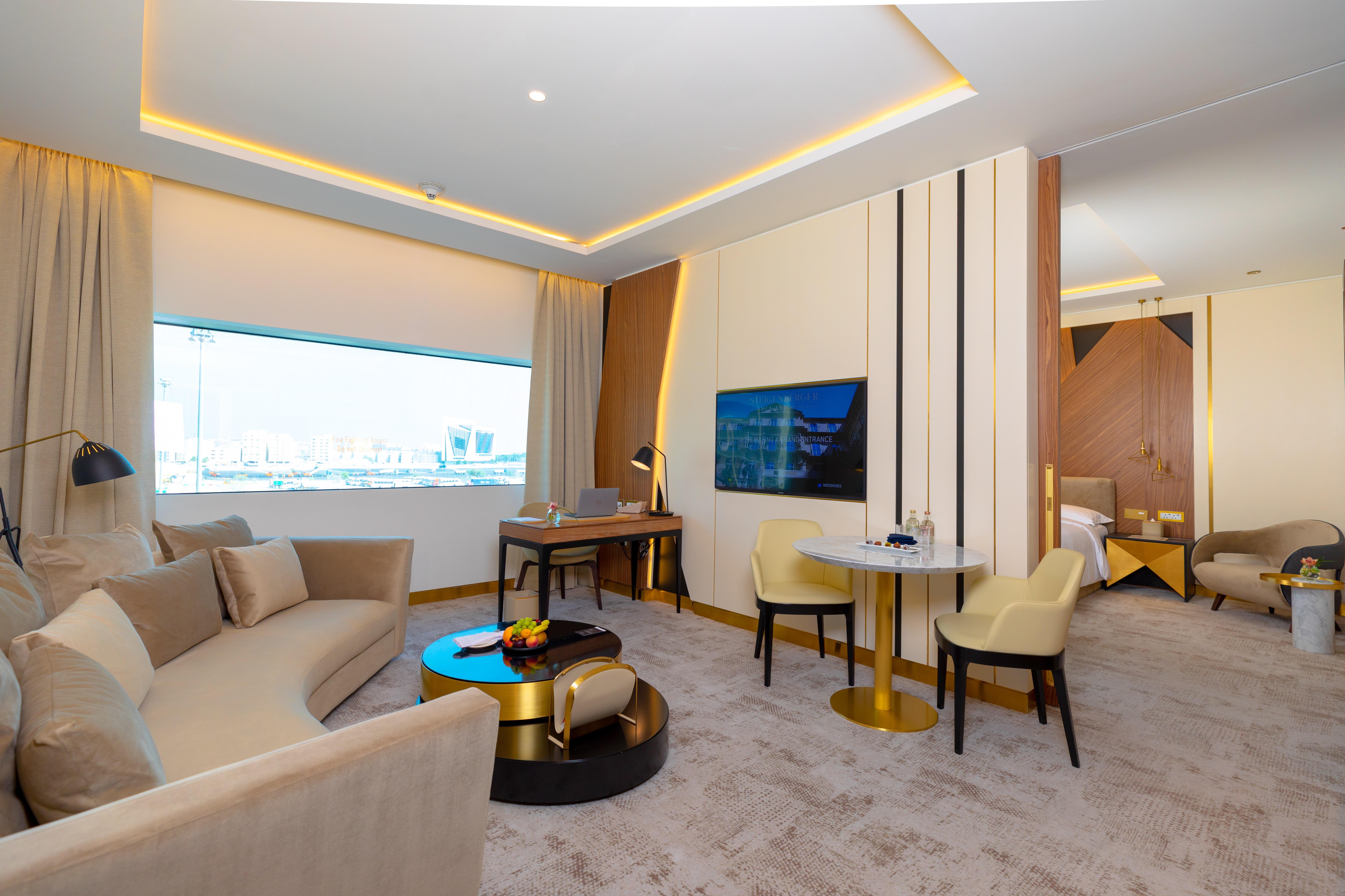 Steigenberger Hotel Доха Екстер'єр фото The photo shows a modern and stylish hotel suite. It features a spacious living area with a curved sofa set and a small round table accompanied by two chairs. There is a large window that allows natural light to fill the room, enhancing its bright an