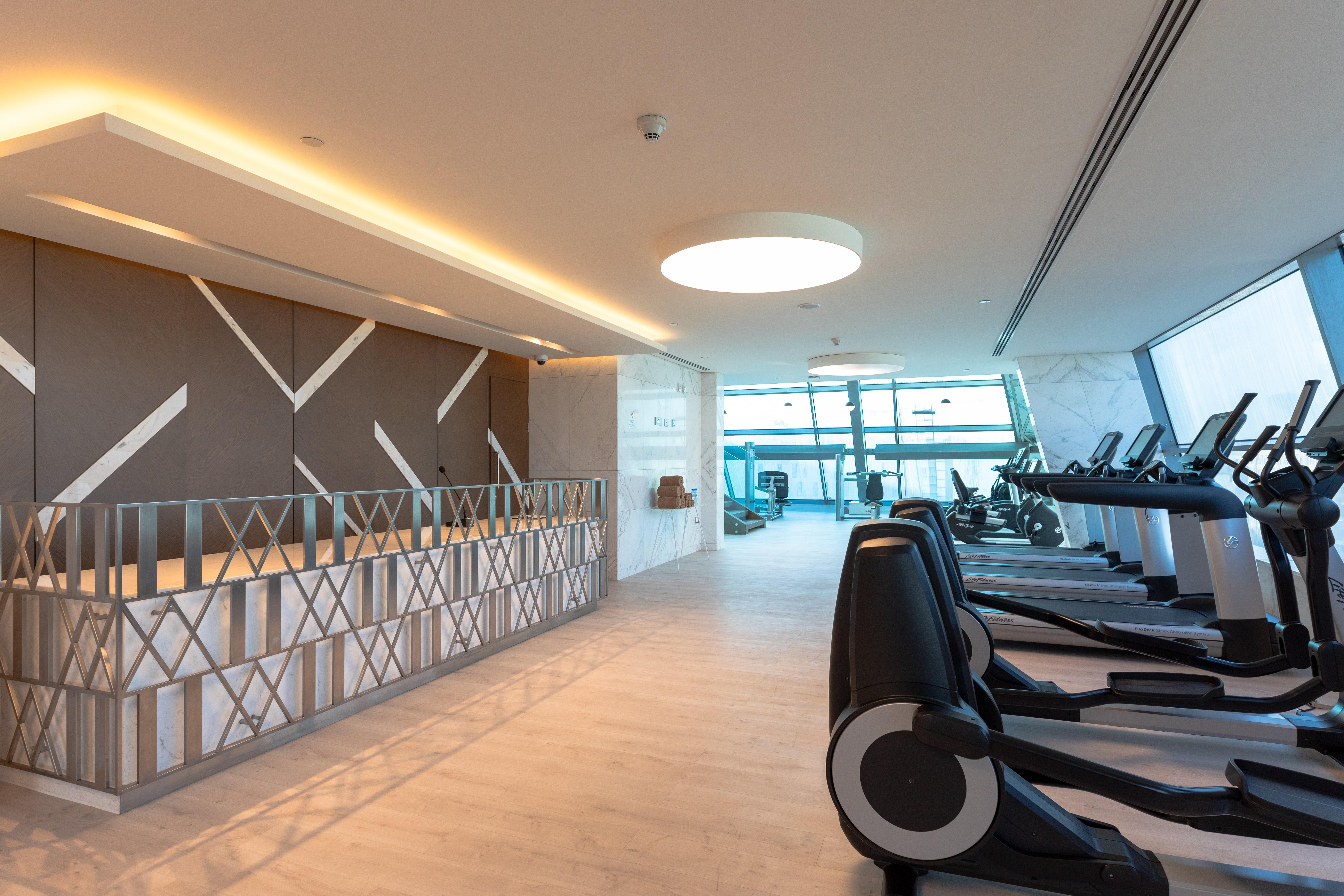 Steigenberger Hotel Доха Екстер'єр фото The photo shows the interior of a modern gym. On the left side, there is a reception area with a stylish design featuring geometric patterns on the wall. The area is well-lit with natural light coming in from large windows, providing a view outside. 