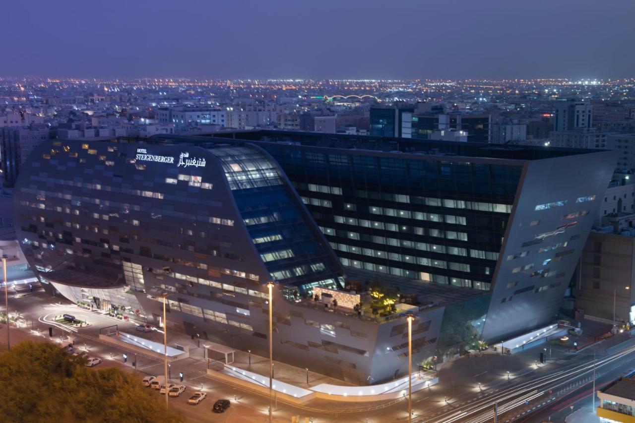 Steigenberger Hotel Доха Екстер'єр фото The image shows a modern architectural building, likely a hotel or office structure, illuminated at night. The design features a unique angular shape with large glass windows, reflecting the surrounding city lights. The building appears to be part of