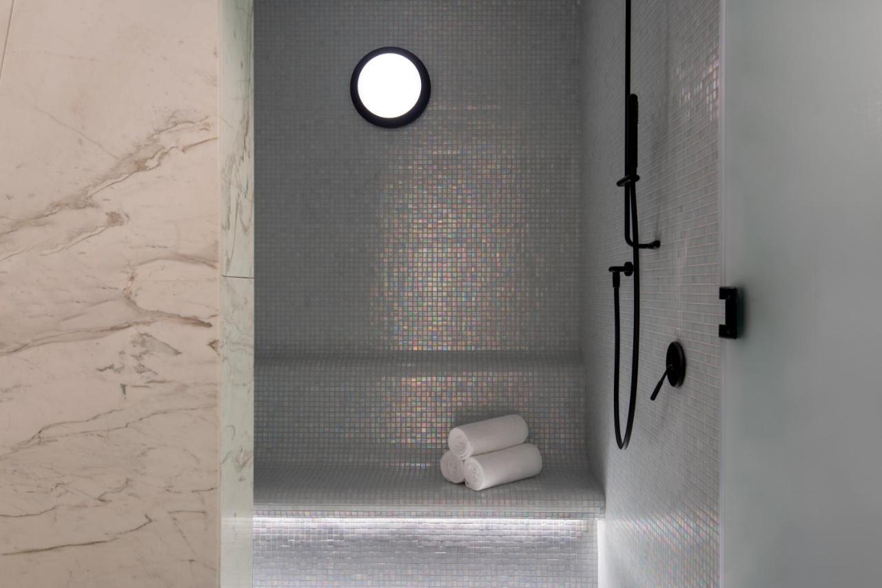 Steigenberger Hotel Доха Екстер'єр фото The photo features a modern shower space. The walls are adorned with small mosaic tiles that create a sleek and reflective surface. There is a round window that allows natural light to enter, enhancing the ambiance. A black shower head and faucet are