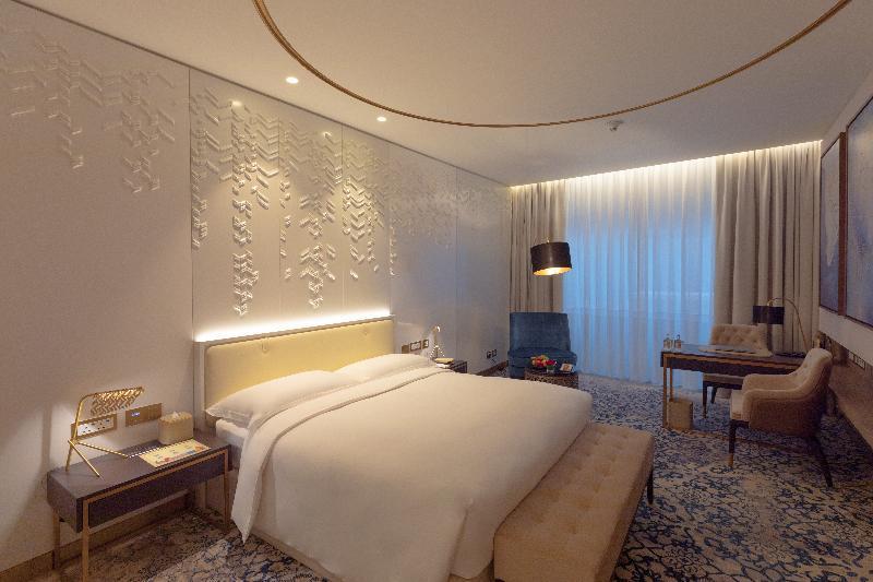 Steigenberger Hotel Доха Екстер'єр фото The photo shows a modern hotel room featuring a contemporary design. The room includes a large bed with a padded headboard, positioned against a textured wall with decorative patterns. To the side, there is a stylish bedside table with a lamp. A comf
