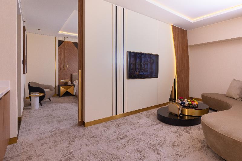 Steigenberger Hotel Доха Екстер'єр фото The photo shows a modern and stylish hotel room or lounge area. The space features a light-colored carpet and walls, with a sleek design. There's a flat-screen TV mounted against a white wall, which has accents of wood and gold. In the foreground, th